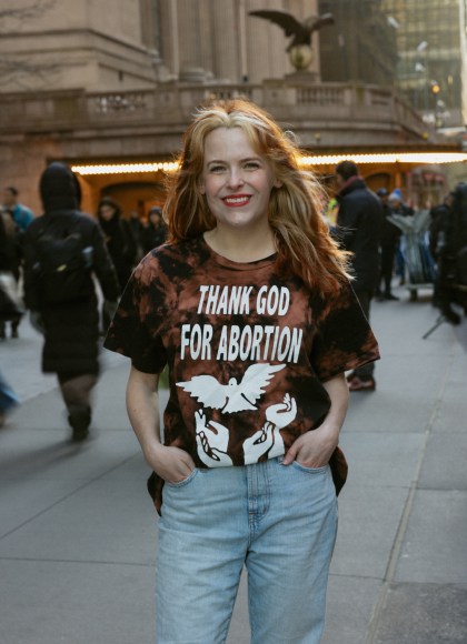 Photo of Becca Rea-Tucker standing on a NYC sidewalk wearing a shirt that reads "Thank God For Abortion" over images of upraised hands and a dove. Becca is smiling at the camera.